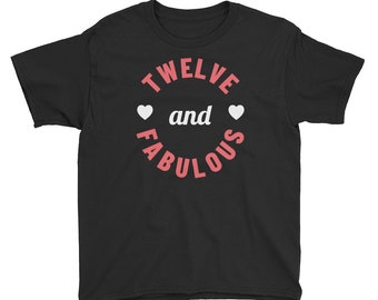 Puede incluir: Camiseta negra para jóvenes con un gráfico circular blanco que dice "TWELVE and FABULOUS" en letras rosas con dos corazones blancos.
