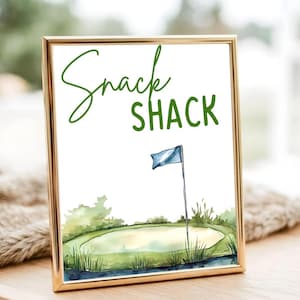 以下が含まれることがあります： 金色のフレームに入ったプリント。水彩画のゴルフコースのシーンで、「Snack Shack」の文字が緑色で描かれています。青い旗も見えます。