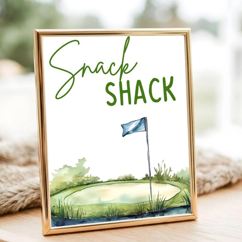 以下が含まれることがあります： 金色のフレームに入ったプリント。水彩画のゴルフコースのシーンで、「Snack Shack」の文字が緑色で描かれています。青い旗も見えます。
