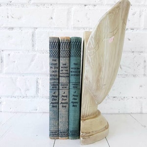 May include: Three vintage Nancy Drew mystery books with blue and green covers. The books are titled "The Clue in the Old Stage Coach", "The Secret of the Golden Pavilion", and "The Hidden Window Mystery". The books are standing on a white surface with a decorative wooden bookend.