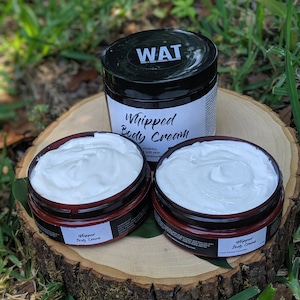 May include: Three jars of whipped body cream, two are brown with white lids and the third is black with a white lid. The jars are on a wooden surface with green grass in the background. The jars are labeled "Whipped Body Cream".