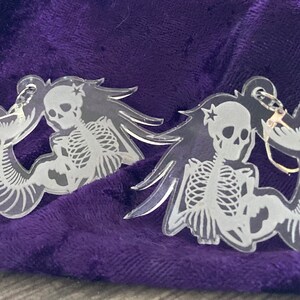 May include: Clear acrylic earrings featuring a skeletal mermaid design. The earrings depict a mermaid skeleton with a star on the head and a fish tail. The earrings are on a purple velvet surface.