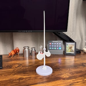 Puede incluir: Un soporte decorativo blanco y gris con un diseño de nube, que sostiene una varilla larga y delgada de color blanco. El soporte está sobre una superficie de madera, con otros objetos en el fondo.
