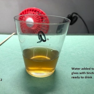 May include: A clear glass shot glass with a brown liquid inside. The glass is labeled with the letter "Q". Water has been added to the shot glass, and it is ready to drink.