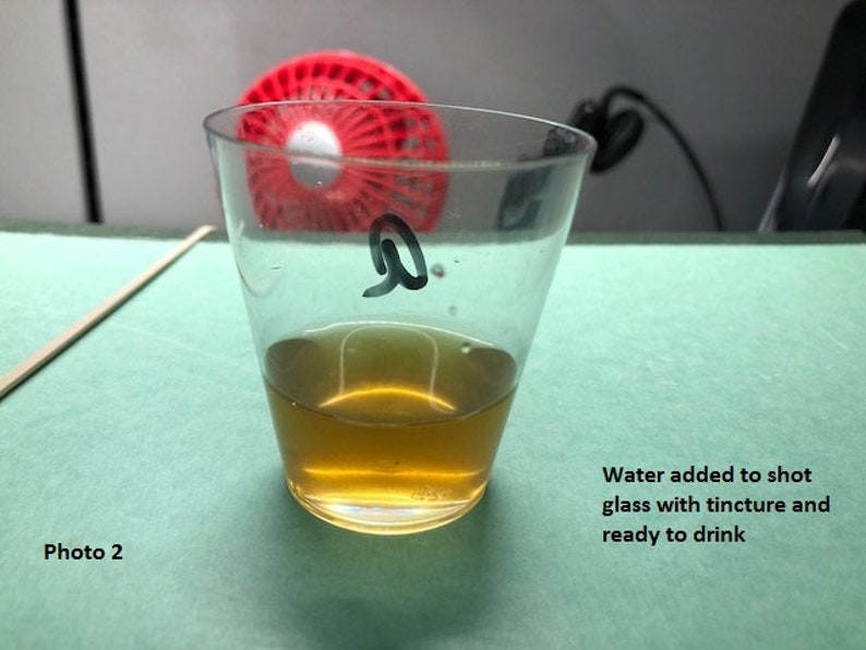 May include: A clear glass shot glass with a brown liquid inside. The glass is labeled with the letter "Q". Water has been added to the shot glass, and it is ready to drink.