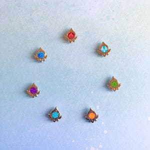 May include: Seven small, gold-colored pins arranged in a circle. Each pin features a unique design with a different color: red, blue, purple, turquoise, orange, green, and teal. The pins have a floral-like base and a teardrop-shaped top.