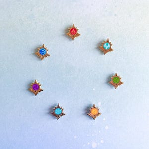 Puede incluir: Siete pines dorados en forma de estrella con centros de diferentes colores. Los centros son morado, azul, rojo, verde, amarillo, naranja y azul claro.