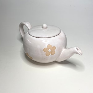 May include: White ceramic teapot with a lid and a handle. The teapot has a floral design with yellow flowers.