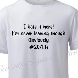 May include: White t-shirt with black text that reads "I hate it here! I'm never leaving though. Obviously. #207life". The shirt has short sleeves and a crew neck.