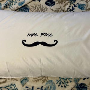 May include: White pillowcase with the text "MRS. ROSS" above a black mustache graphic. The pillowcase is rectangular and appears to be made of a smooth fabric. The pillowcase is laying on a patterned blanket.