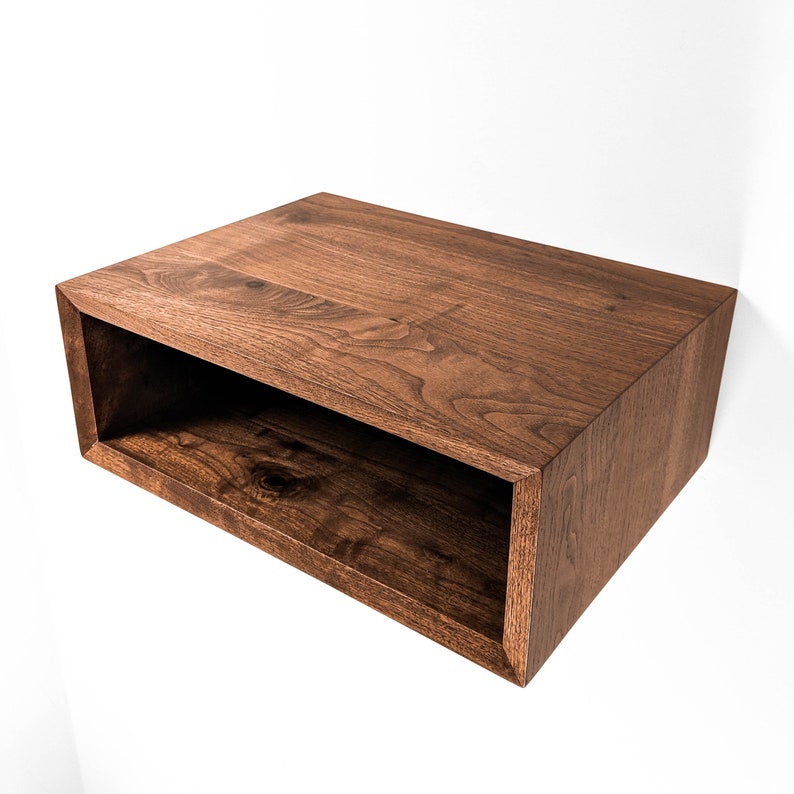 May include: A rectangular, open-front floating shelf made of dark brown wood. The shelf is mounted on a white wall, showcasing the wood grain and the clean lines of the design. The shelf is approximately 24 inches wide.