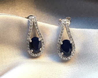 May include: A pair of silver-toned stud earrings. Each earring features a dark blue oval gemstone surrounded by small, clear stones. The earrings are set against a white, silky background.