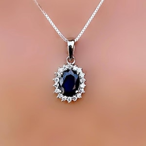 Könnte beinhalten: Eine silberne Halskette mit Anhänger, die einen ovalen, dunkelblauen Edelstein zeigt, der von kleinen, klaren Edelsteinen umgeben ist. Der Anhänger hängt an einer zarten Silberkette. Der Schmuck wird vor einem weichen, neutralen Hintergrund präsentiert.