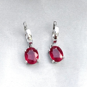 Könnte beinhalten: Ein Paar silberne Creolen mit baumelnden roten ovalen Edelsteinen.