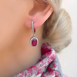 May include: A pair of silver hoop earrings with a dangling oval ruby gemstone surrounded by a halo of smaller diamonds.