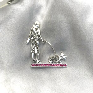 Op de afbeelding: Een zilveren broche met een vrouw die een hond uitlaat. Beide figuren zijn gemaakt van marcasiet en zijn geplaatst op een basis van roze stenen.