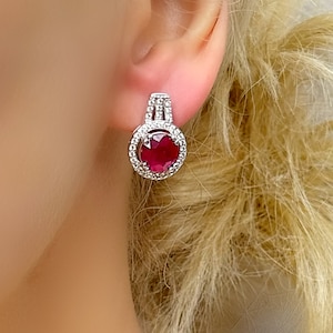 May include: Silver-coloured earrings featuring a large, round, deep red gemstone. The gemstone is encircled by smaller, clear stones. The earrings are set against a blurred background.