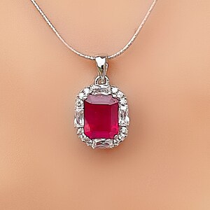 May include: A silver chain necklace with a pendant featuring a large, red, square-shaped gemstone surrounded by smaller, clear gemstones.