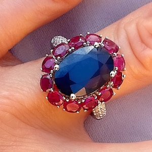 May include: A ring featuring a large oval blue sapphire, surrounded by a halo of red rubies and small diamonds.