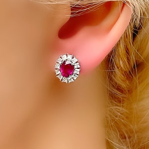 May include: A pair of silver stud earrings with a red oval ruby gemstone surrounded by a halo of smaller white gemstones.