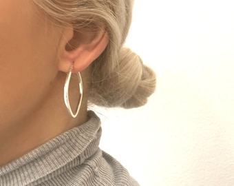 May include: Silver hoop earrings with a unique, slightly teardrop-shaped design. The earrings are shown on a model with blonde hair.