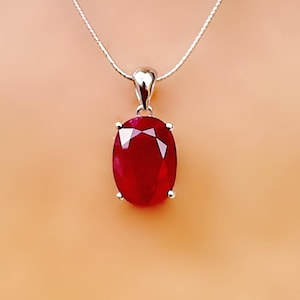 Könnte beinhalten: Eine silberne Halskette mit einem großen, oval geschliffenen Rubinanhänger. Der Rubin hat eine tiefe rote Farbe und ist in einer silbernen Lünette gefasst. Die Halskette ist schlicht und elegant, geeignet für den Alltag oder besondere Anlässe.