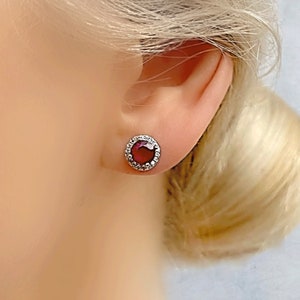 Peut inclure: Gros plan d'une boucle d'oreille à clou avec une pierre précieuse rouge foncé ronde, entourée d'un halo de petites pierres claires. La boucle d'oreille est sertie dans un métal argenté. La boucle d'oreille est sur l'oreille d'une personne.
