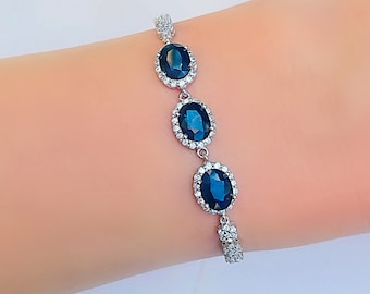Op de afbeelding: Een zilveren armband met drie ovale, donkerblauwe edelstenen, elk omringd door kleinere, heldere stenen. De armband wordt getoond op een arm, wat het elegante ontwerp benadrukt.