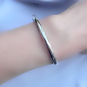 Peut inclure: Un bracelet jonc argenté avec une finition polie. Le bracelet a un design simple et élégant, reflétant la lumière. Le bijou est porté sur un poignet sur un fond bleu clair.