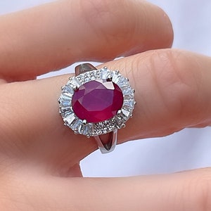 May include: A silver ring featuring a large oval ruby gemstone surrounded by smaller white diamonds.