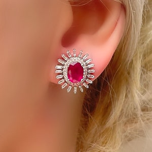 Op de afbeelding: Een paar zilveren oorbellen met een grote ovale rode edelsteen omringd door kleinere heldere edelstenen.