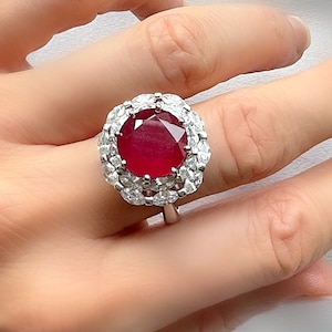 May include: A silver ring with a large, round, red ruby gemstone surrounded by a halo of smaller, clear gemstones.