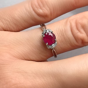 May include: A silver ring with a striking red oval gemstone, encircled by a halo of smaller, clear stones. The ring's delicate band highlights the central stone. This elegant piece of jewellery is a classic and eye-catching accessory.