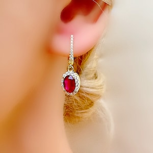 Puede incluir: Un pendiente dorado con una vibrante gema roja de forma ovalada, rodeada por un halo de pequeñas piedras blancas brillantes. El pendiente tiene una sección superior delgada y adornada, creando un elegante diseño colgante. La joya se encuentra sobre un fondo suave y borroso.