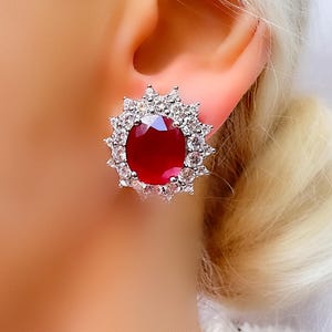 May include: A silver stud earring featuring a large, oval, deep red gemstone surrounded by a halo of smaller, clear gemstones. The design resembles a sunburst. The earring is set against a blurred background.