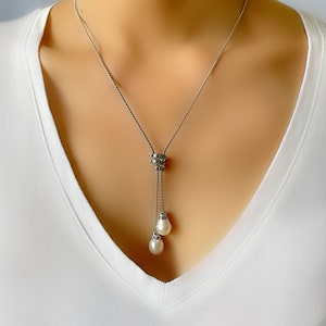 May include: A silver-toned necklace with a delicate chain and a pendant featuring two teardrop-shaped pearls. The necklace has a decorative element at the top of the pendant, adding a touch of elegance. The necklace is worn against a white v-neck shirt.