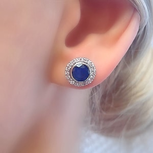 May include: A pair of silver stud earrings featuring a round blue gemstone encircled by a halo of smaller white gemstones.