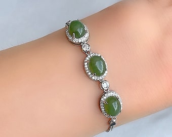 Op de afbeelding: Een zilveren armband met drie ovale, groene edelstenen. Elke edelsteen is omgeven door een halo van kleine, heldere kristallen. De armband wordt getoond op een pols.