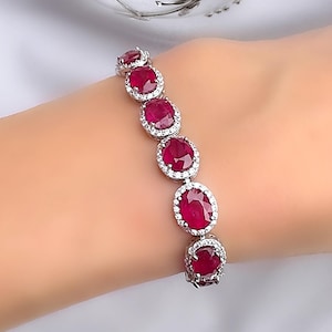 Könnte beinhalten: Ein silbernes Armband mit ovalen, tiefroten Edelsteinen, die jeweils von kleineren, klaren Steinen umgeben sind. Das Armband wird an einem Handgelenk vor weißem Hintergrund gezeigt, um das Funkeln und Design des Schmucks hervorzuheben.