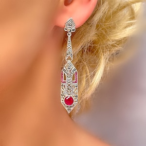 May include: A pair of silver dangle earrings with a red gemstone and marcasite accents. The earrings are in an art deco style.