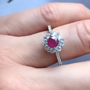 Op de afbeelding: Een zilveren ring met een rode robijn-edelsteen omringd door kleinere witte edelstenen. De ring is gezet in een bloemvormig ontwerp.