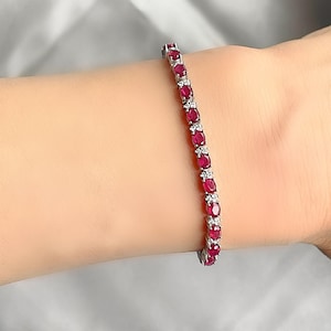 May include: A tennis bracelet featuring a row of oval-shaped red rubies set in a silver metal band.