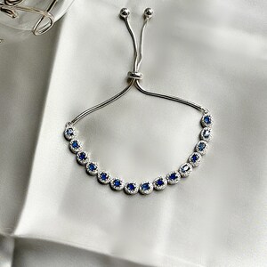 May include: Silver-toned bracelet featuring a row of oval, deep blue gemstones, each surrounded by smaller, clear stones. The bracelet has an adjustable sliding clasp and is displayed on a white, silky surface. A piece of jewelry.