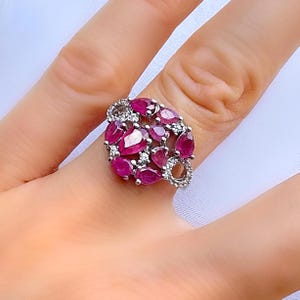 May include: A silver ring featuring a cluster of oval and pear-shaped rubies. Small, round diamonds are interspersed among the rubies. The ring's design is a floral or abstract arrangement, with a bright, eye-catching appearance.
