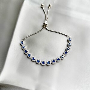 May include: A silver bracelet with a row of oval, deep blue gemstones, each surrounded by smaller, clear stones. The bracelet has a sliding clasp for adjustable sizing. The background is a soft, white fabric.