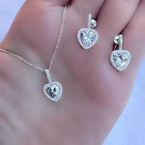 Könnte beinhalten: Ein silberfarbenes Schmuckset mit einer herzförmigen Anhängerkette und passenden Ohrringen. Jedes Schmuckstück ist mit einem großen, klaren, herzförmigen Stein verziert, der von kleineren, funkelnden Steinen umgeben ist. Die Ohrringe haben einen Steckerverschluss.
