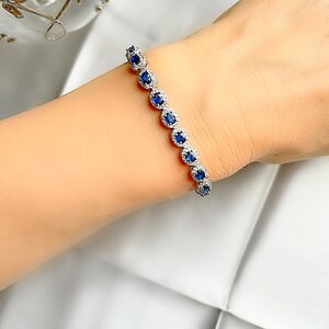 May include: A silver bracelet with oval, deep blue gemstones encircled by smaller, clear stones. The bracelet is displayed on a wrist against a white background. The gemstones are evenly spaced along the bracelet's length.
