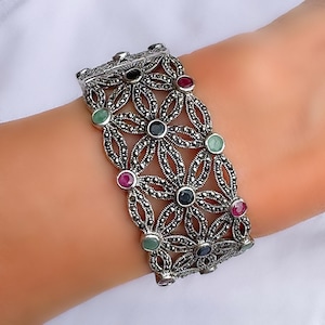 Puede incluir: Una pulsera ancha de plata con un intrincado patrón floral calado incrustado con pequeñas piedras de marcasita gris oscuro. El diseño está acentuado con piedras preciosas cabujón redondas y pulidas en tonos rojo intenso, azul oscuro y verde claro.