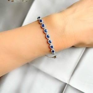 May include: A silver bracelet featuring a series of round, dark blue gemstones encircled by smaller, clear stones. The bracelet is displayed on a wrist against a soft, white background, showcasing its elegant design and sparkle.