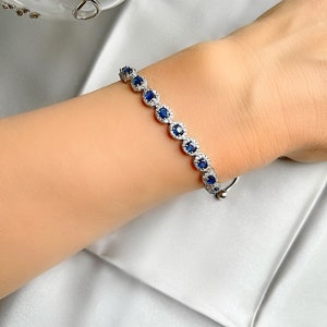 May include: A silver bracelet featuring a series of round, deep blue gemstones encircled by smaller, clear stones. The bracelet is displayed on a wrist against a backdrop of a silky, white fabric.